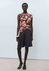 Top floreale a spalla scoperta di colore marrone scuro con motivi rosa e verdi, abbinato a pantaloni corti marroni di lunghezza al ginocchio. Gli accessori includono un bracciale largo.