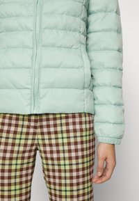 Light teal puffer jacket with horizontal quilted pattern and front zipper. Worn over checked pants in brown, green, and cream colors.