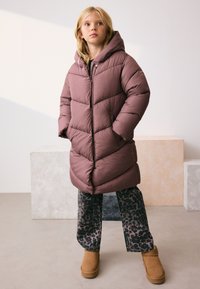 Fioletowy, pikowany płaszcz z kapturem, ozdobiony przeszyciami w jodełkę. Do tego czarne spodnie w panterkę i beżowe botki za kostkę. Gładka faktura.