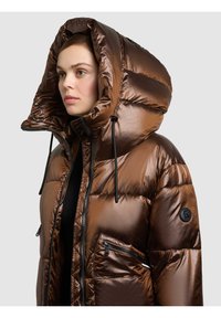 Brązowa puchowa kurtka z dużym, kapturowym kołnierzem, błyszczącego materiału, czarnymi suwakami oraz naszywką na lewej rękawie. Posiada kieszeń na zamek.