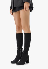 Botas negras hasta la rodilla de ante con tacón cuadrado. Presentan un cierre lateral y un acabado a costura en la parte superior. Textura suave.