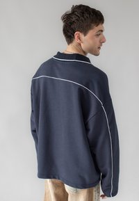 Marinblå oversized tröja med texturerad yta, med vit kantdetalj som framhäver axlarna och en ribbad krage.