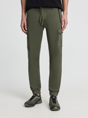 Osoba nosząca oliwkowo-zielone spodnie typu cargo jogger z kieszeniami na zamek błyskawiczny i z patkami, w połączeniu z pasującymi zielono-czarnymi butami sportowymi.