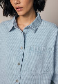 Light blue button-up shirt featuring a textured, striped pattern, a chest pocket, and silver button hardware. Collared design.