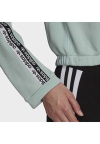 Ljus mintgrön sweatshirt med svart logoband, ribbade ärmar och avslappnad passform, ihop med svarta byxor med vita ränder.