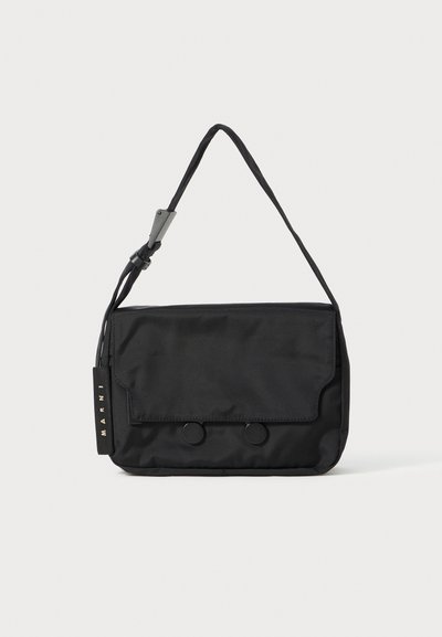 Sac à épaule noir en nylon, de forme plate, avec une fermeture à rabat, deux boutons ronds et une bandoulière tissée. Étiquette de marque visible.