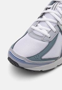 Baskets blanches avec des accents gris et bleus, dessus en mesh respirant, lacets blancs et semelle en caoutchouc texturée visible au niveau des orteils.