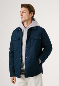 Chaqueta de navidad acolchada en color azul marino con botones a presión y dos bolsillos en el pecho, llevada sobre una sudadera gris con capucha. Cuenta con un ajuste relajado y una textura suave.