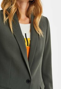 Olivově zelené sako na míru s klopy s výstřihem do V, jedním černým knoflíkem a hladkou texturou; nošené přes oranžové tričko s grafikou.