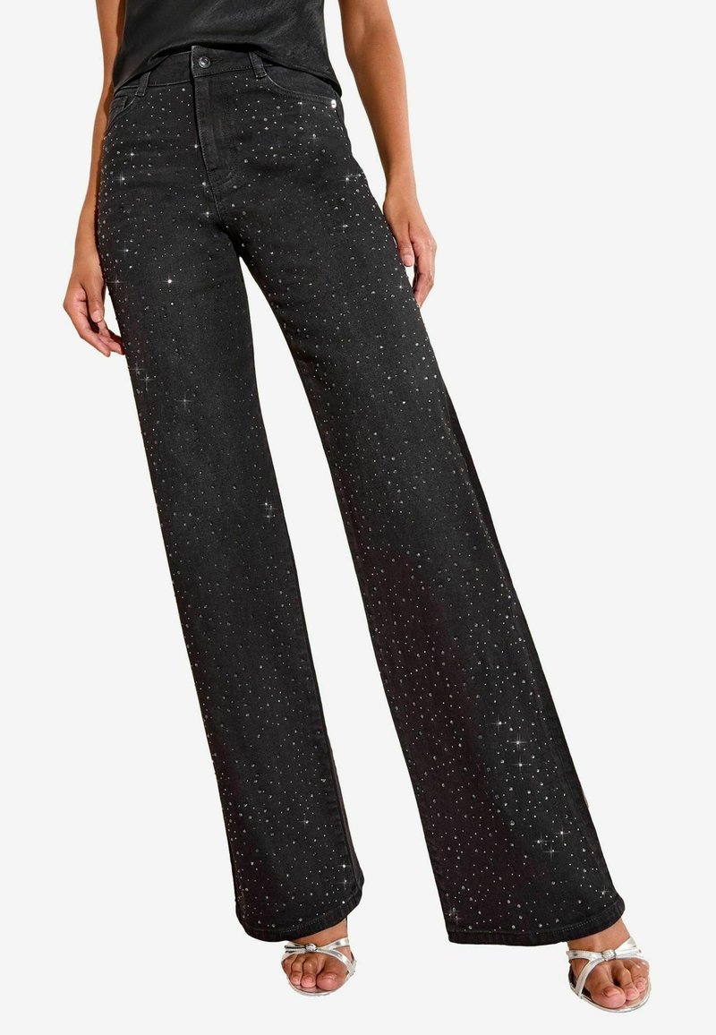 Melni platleģēti džinsi ar izkaisītām sudraba spīdīgām punktiem, valkāti kopā ar sudraba siksniņu atvērtu purngalu sandalēm un melnu topu.