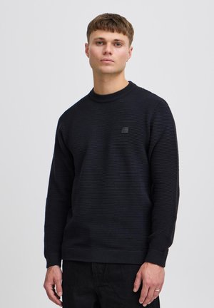 Young man with short hair wearing a black textured long-sleeve sweater and black pants against a plain light background.