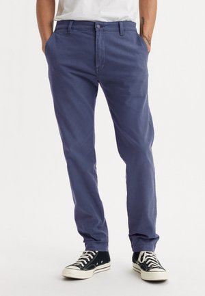 Pantalon chino en coton violet foncé avec une coupe slim, doté de poches latérales et d'une fermeture à bouton à l'avant, associé à des baskets en toile noires.