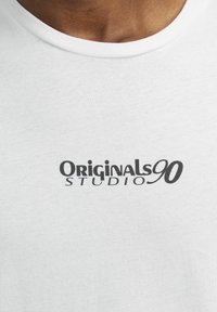 Bílý bavlněný tričko s kulatým výstřihem, na kterém je černé logo s nápisem "Originals90 STUDIO" v tučné typografii.