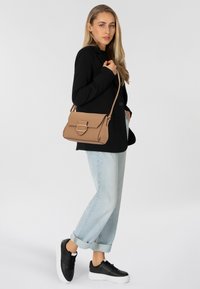 Borsa strutturata color tan con forma rettangolare e un importante gancio metallico, abbinata a un blazer nero e pantaloni di jeans chiari arrotolati.