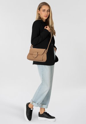 Beige strukturierte Handtasche mit rechteckiger Form und einem auffälligen metallischen Verschluss, kombiniert mit einem schwarzen Blazer und hellen, gerollten Jeanshosen.