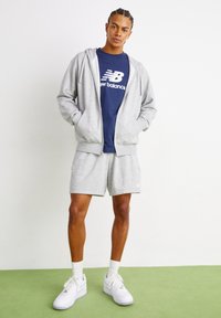 Vyras, apsirengęs pilka užtraukiama striuke, derančiomis pilkomis šortais, mėlynu "New Balance" marškinėliais, baltais kojiniais ir baltais sportiniais batais, stovi ant žalios grindų.