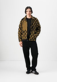 Brown and black patterned fleece jacket with zip closure, front pockets, paired with a black t-shirt and black jeans. Black sneakers.