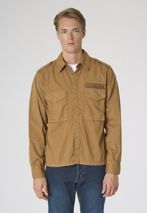 Giovane uomo che indossa una camicia color cuoio con bottoni e toppa "U.S. Polo Assn.", e jeans blu, in piedi davanti a uno sfondo semplice.