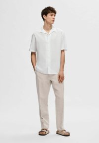 Camicia bianca a maniche corte con bottoni, pantaloni di lino beige e sandali marroni su un modello maschile in piedi contro uno sfondo semplice.