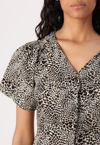 Robe à manches courtes en tissu beige avec un imprimé de cœurs noirs et marron, col en V et boutonnage sur le devant. Texture lisse avec un léger éclat.