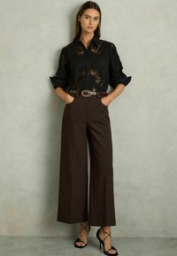 Zwarte kant blouse met knoopsluiting aan de voorkant, gecombineerd met bruin hoge taille brede broek en zwarte schouderhaksandalen.
