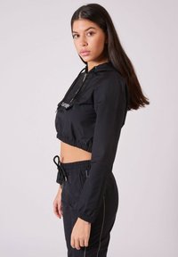 Schwarze, cropped Jacke mit Kapuze, die über eine Fronttasche, einen Kordelzug und einen elastischen Saum verfügt. Kombiniert mit passenden schwarzen Jogginghosen.
