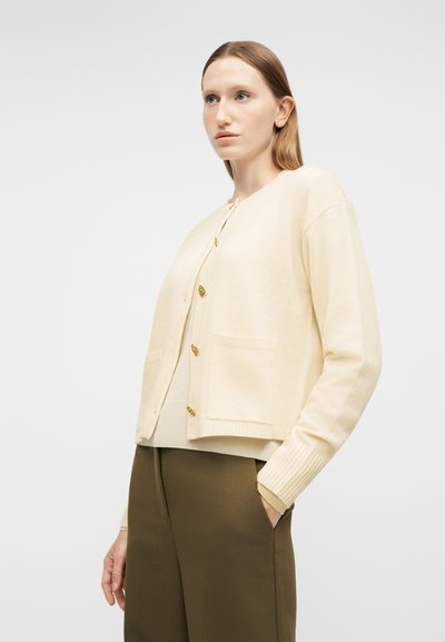 Γυναίκα με ίσια ξανθά μαλλιά που φοράει εκρου ζακέτα με κουμπιά και ελαιδί παντελόνι, στέκεται με το ένα χέρι στην τσέπη μπροστά σε λευκό φόντο.