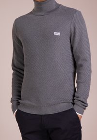 Pull à col roulé gris en tissu tricoté texturé, présentant un motif chevrons subtil et un petit logo blanc sur la poitrine.