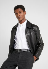 Veste en cuir noir avec une fermeture éclair à l'avant, un grand col et des poches latérales, portée par-dessus une chemise blanche à boutons et un pantalon gris.