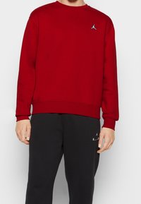 Sudadera roja de manga larga, con cuello redondo y bordes acanalados. Presenta un pequeño logo azul en el pecho, combinado con pantalones de chándal oscuros.