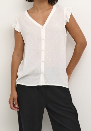 Persoon die een witte mouwloze blouse met knopen draagt, met getextureerde stippen en geplooide schouders, gecombineerd met losse zwarte broek tegen een effen achtergrond.