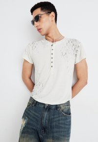 Kortärmad, ljus krämfärgad t-shirt med ormtryck på ena sidan och femknappsslå, kombinerad med urtvättade blå jeans.