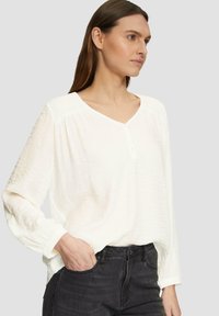 Blouse blanche en tissu texturé, avec un décolleté en V et des détails de boutons, à manches longues et à coupe décontractée. Associée à un jean foncé.