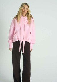 Růžová mikina s kapucí, přední kapsou, stahovací šňůrkou a vyšitým nápisem. Kombinována s širokými tmavě hnědými kalhotami. Měkká, neformální látka.