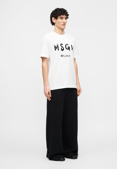Jeune homme debout, portant un t-shirt blanc MSGM Milano et un pantalon noir à jambes larges, légèrement tourné vers la droite sur un fond blanc uni.