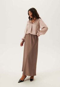 Lunga gonna marrone abbinata a una blusa di seta rosa chiaro con scollatura rotonda e maniche a sbuffo. La modella è in piedi su uno sfondo neutro.