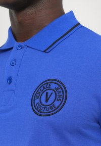 Μπλε πόλο μπ�λούζα με υφή υφάσματος, που διαθέτει μαύρη ριγέ κολάρο, τρία κουμπιά και κεντητό κυκλικό λογότυπο σε μαύρο και άσπρο.