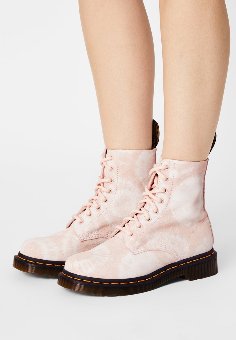 Personne portant des bottines à lacets tie-dye rose clair, avec des semelles épaisses noires et des coutures jaunes, debout sur une surface blanche.