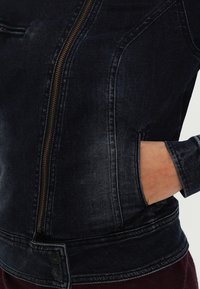 Džínová bunda v tmavě modré barvě, sázkovým zlatým zipem, bočními kapsami a knoflíkovými detaily na lemu. Textura je hladká a mírně opotřebovaná.