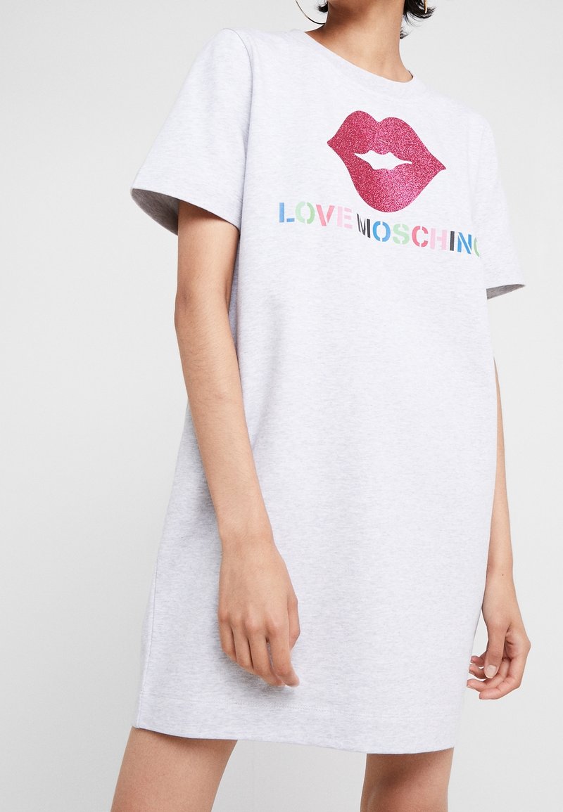 Kvinna i en ljusgrå kortärmad klänning med glittriga rosa läppar och flerfärgad text "LOVE MOSCHINO" på framsidan.