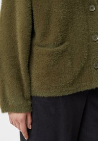 Oliwkowo zielony, puszysty kardigan z pojedynczą kieszenią, okrągłymi guzikami i fakturowaną tkaniną. Rękawy są lekko oversize.
