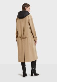 Beige trench coat med en svart pufferhuva, åtsittande midja med bälte och långa ärmar. Slät tyg med en lös passform och minimal design.