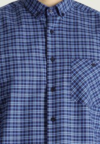 Camicia a quadretti nei toni navy e azzurro chiaro, con colletto, bottoni frontali e tasca sul petto con un motivo diagonale.