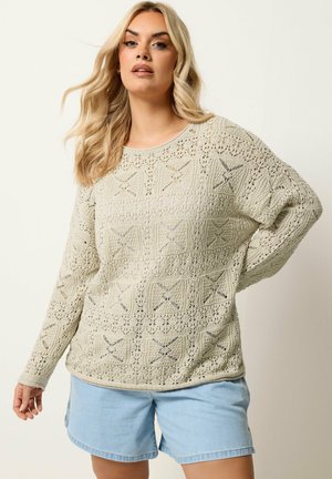 Blonde Frau trägt einen cremefarbenen langärmeligen Häkelpullover und hellblaue Jeansshorts, posiert mit einer Hand auf der Hüfte vor einem einfarbigen Hintergrund.