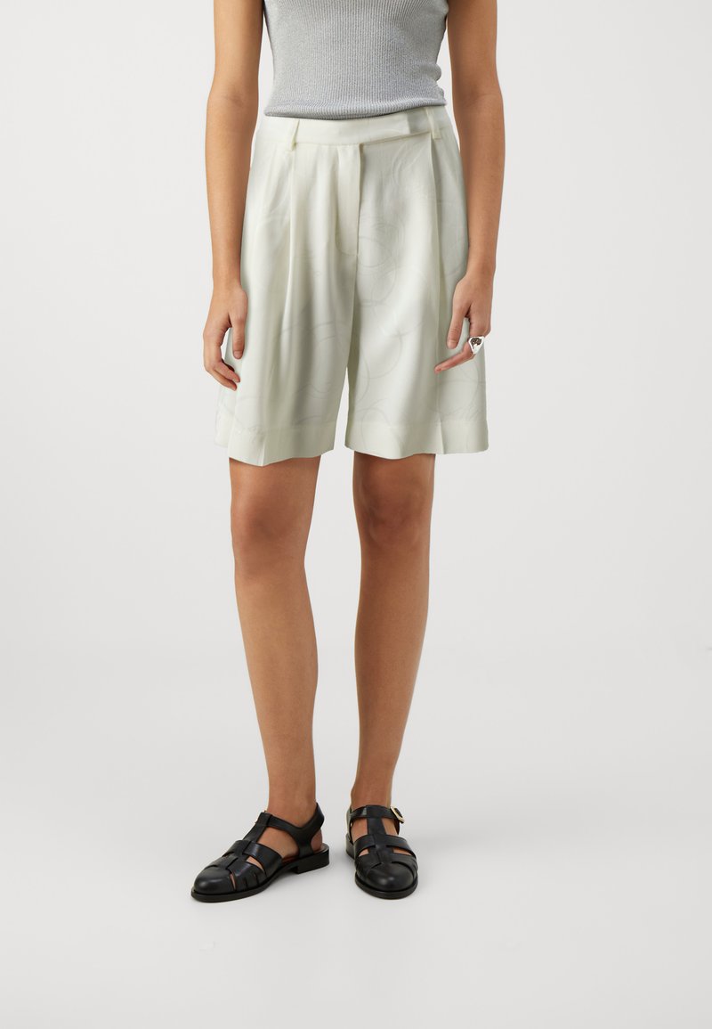 Light green pleated shorts made of smooth fabric, featuring a subtle pattern. Paired with black strappy sandals for contrast.