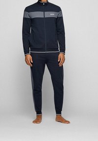 Marinträningsdräkt som består av en zip-up jacka med grå horisontella ränder och en vit logotyp, i kombination med tapered byxor. Tillverkad i ett mjukt tyg.