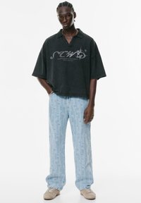 Jonge man met gevlochten haar, gekleed in een losse zwarte grafische polo, lichtblauwe gebloemde jeans, beige sneakers en zilveren sieraden.