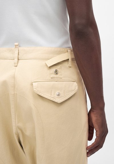 Gros plan d'une personne portant un pantalon beige avec une poche arrière boutonnée et une patte latérale réglable, associé à un haut blanc côtelé.
