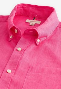 Chemise rose vif boutonnée avec col boutonné et poche poitrine posée à plat, montrant la texture du tissu et l'étiquette de la marque à l'intérieur du col.