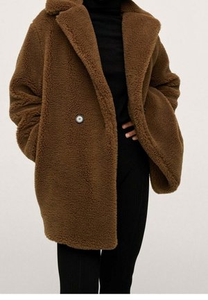 Manteau teddy marron avec grands revers et boutons porté sur un pantalon côtelé noir et un haut noir, mains dans les poches du manteau.